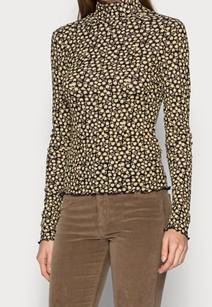 Nainen pukeutunut istuvaan mustaan poolopaitaan, jossa on pieni beige kukkakuosi, ja ruskeisiin corduroy-housuihin.