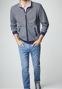 Chaqueta con cremallera en navy con detalles en gris, que presenta dos bolsillos frontales y puños acanalados, combinada con jeans azul claro y zapatillas grises.