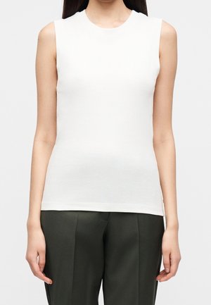 Femme portant un haut blanc sans manches et un pantalon vert foncé, debout avec les bras détendus le long du corps sur un fond uni.