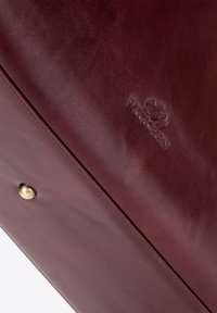 Bordeaux leren tas met een gladde textuur, voorzien van een metalen drukknoopsluiting en een bedrukt logo, die een minimalistisch ontwerp en klassieke aantrekkingskracht toont.