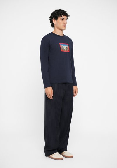 Navy long-sleeve shirt with a rectangular photo print and red outline, paired with black pinstriped trousers and light-colored shoes.