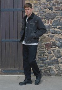 Melna džinsu jaka ar aitas vilnas apkakli, balta krekls, tumšie bikses un melni zābaki, fona ir akmens siena un koka durvis.