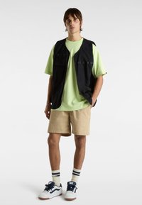 Black utility vest over a light green t-shirt, paired with beige shorts. White sneakers with navy accents and striped socks. Simple design.