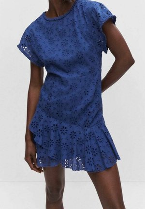 Femme portant une robe bleue à manches courtes avec motif floral ajouré et un ourlet asymétrique à volants, debout, une main dans le dos.