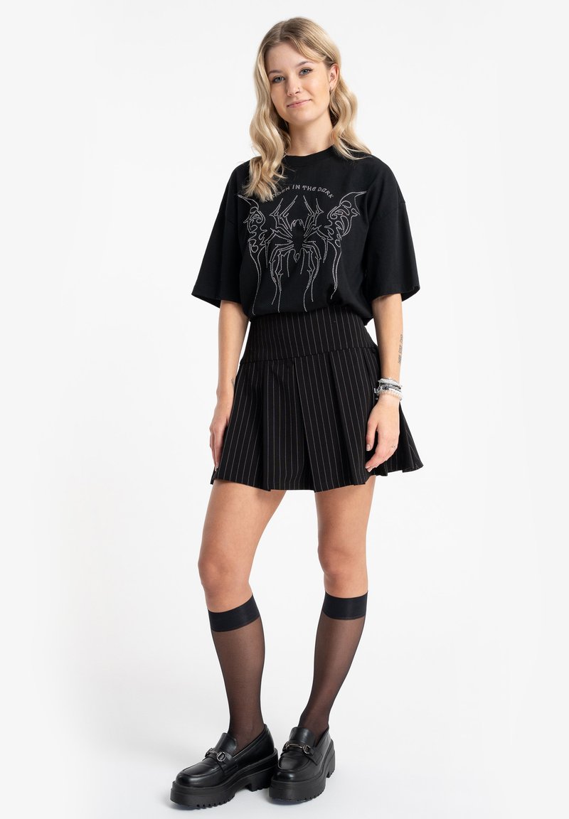 Czarny, oversize'owy t-shirt z graficznym wzorem, zestawiony z czarną plisowaną spódnicą w cienkie prążki. Modelka nosi przezroczyste czarne skarpetki do kolan oraz masywne buty.