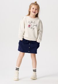 Hellbeige Sweatshirt mit dem Aufdruck "Was für ein schöner Tag" und einer pinken Blume, kombiniert mit einem marineblauen Rock mit Knöpfen und seitlichen Taschen.