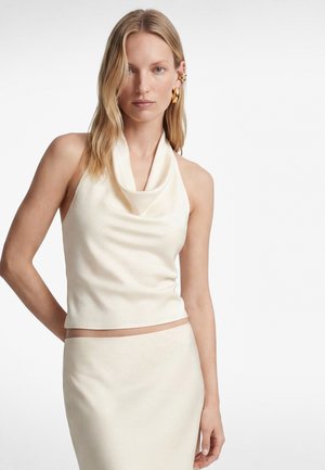 Blonde vrouw met een crèmekleurige haltertop met gedrapeerde halslijn en bijpassende rok, staand tegen een witte achtergrond.
