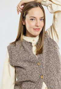 Jonge vrouw met steil bruin haar, gekleed in een crème knoopshirt en een bruine gebreide vest, die één arm opsteekt en zachtjes glimlacht.