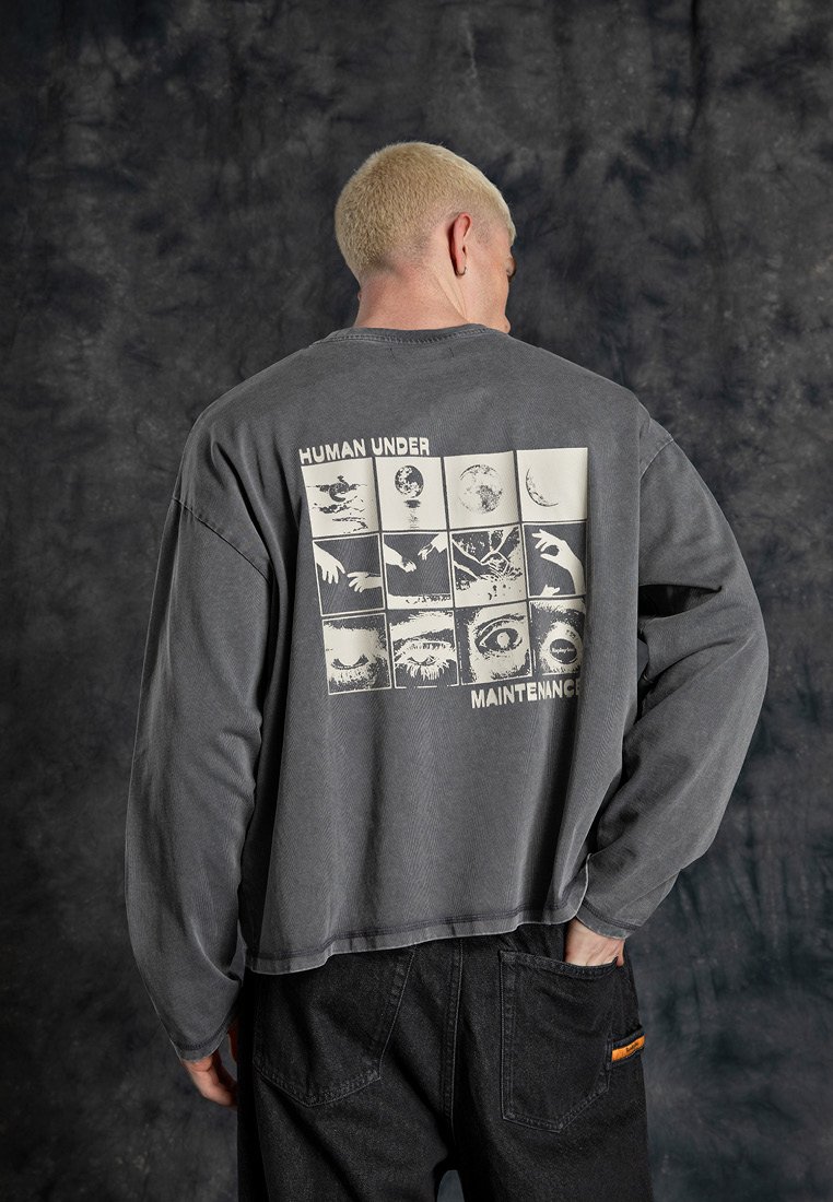 Grå långärmad t-shirt i bomull, med en grafisk design på baksidan som visar fyrkanter med olika bilder och texten "HUMAN UNDER MAINTENANCE."