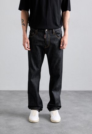 Man wearing black jeans with visible yellow stitching, white sneakers, black shirt, and tattoos on forearms against plain gray background.
