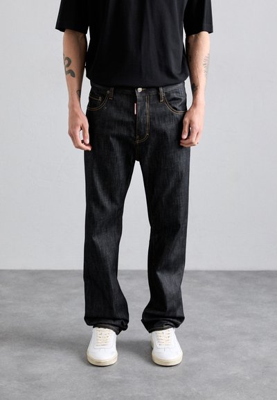 Man wearing black jeans with visible yellow stitching, white sneakers, black shirt, and tattoos on forearms against plain gray background.