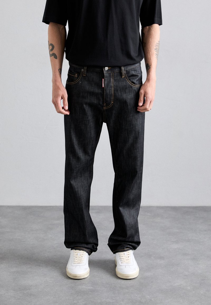 Homme portant un jean noir avec des coutures jaunes visibles, des baskets blanches, un shirt noir et des tatouages sur les avant-bras, sur fond gris uni.