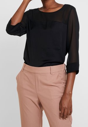 Mujer con una blusa negra transparente de manga larga metida dentro de unos pantalones beige de cintura alta, con las manos relajadas a los lados.