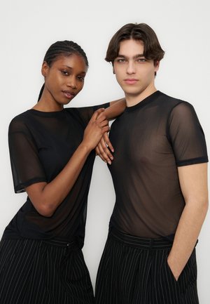 Dvaja mladí dospelí, oblečení v čiernych priesvitných topoch a čiernych nohaviciach s tenkými pruhmi, stojaci blízko seba, pričom jeden si opiera ruku o rameno toho druhého na bielom pozadí.