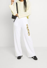 Sudadera corta amarilla con cordones, combinada con pantalones anchos blancos que presentan bordados de girasoles a lo largo del costado, complementados con zapatillas negras.