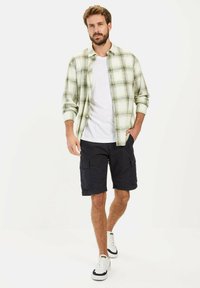 Kariertes Herrenhemd mit Knopfleiste in Hellgrün und Weiß, kombiniert mit schwarzen Cargo-Shorts und weißen Sneakern mit schwarzen Akzenten.