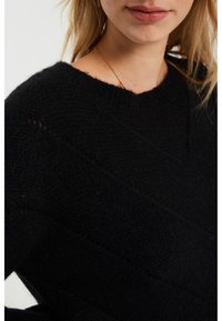 Černý pletený svetr s kulatým výstřihem, texturovaným vzorem a mírně spuštěnými rameny. Látka působí měkce a teple.