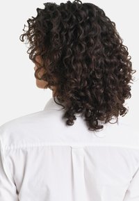 Lockige, dunkle Haare fallen über die Schultern, dazu ein weißes, knöpfbares Hemd mit einer Passe und subtiler Faltentiefe am Rücken.