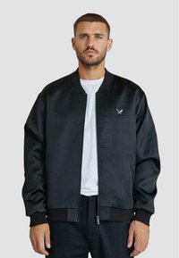 Giacca bomber nera realizzata in tessuto morbido, con colletto e polsini a coste. Include un piccolo emblema con spade incrociate sul petto.