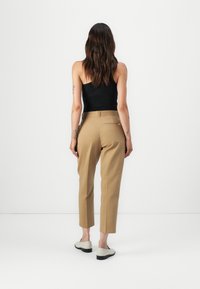 Pantaloni beige su misura con vita alta, dotati di una tasca posteriore e taglio dritto, abbinati a un top senza maniche nero e scarpe chiare.