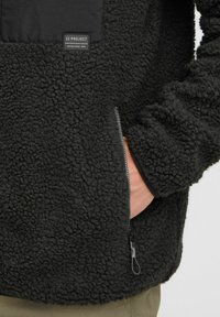 Veste en polaire noire dotée d'une surface texturée, d'une poche zipée sur le côté et d'une étiquette en tissu noir sur la poitrine avec un logo blanc.