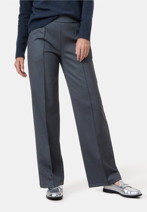 Vrouw draagt donker textuurrijke wijde broekspijpen, marineblauwe trui en zilveren loafers, staat met één hand ontspannen langs het lichaam.