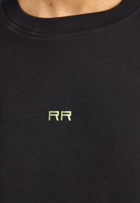 Camiseta negra de algodón con cuello redondo y un pequeño logo reflectante amarillo "RR" en el pecho. Textura suave, diseño minimalista.