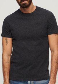 T-shirt in cotone grigio scuro con scollo rotondo, maniche corte e un piccolo logo ricamato sul petto. Texture uniforme con dettagli minimi.