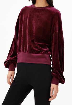 Femme portant un haut ample en velours bordeaux à manches longues avec des manches retroussées et une large ceinture, associé à un pantalon noir.