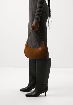 Personne en robe gris côtelée tenant un sac à main marron en forme de croissant, portant des bottes noires à talons hauts, à bout pointu, montantes jusqu'aux genoux, debout sur un fond blanc.