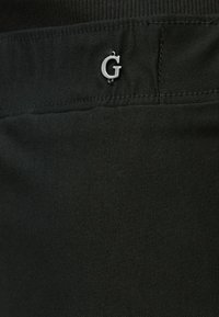 Μαύρο ύφασμα με λεία υφή, που διαθέτει ασημένιο λογότυπο "G" ως διακριτικό στοιχείο στην πίσω τσέπη, αναδεικνύοντας μινιμαλιστικά στοιχεία σχεδίασης.