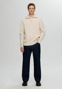 Suéter texturizado de color crema con cuello de media cremallera, combinado con jeans rectos de color azul oscuro y zapatos negros.