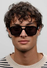 Zwarte rechthoekige zonnebril met donkere lenzen, zichtbaar is een gepolijst montuur. Het model draagt een gestreepte trui, met krullend haar zichtbaar.