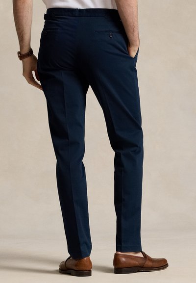 Άνδρας που φοράει ναυτικά ραμμένο παντελόνι και καφέ δερμάτινα μοκασίνια, στέκεται με το ένα χέρι στην τσέπη σε μπεζ φόντο.