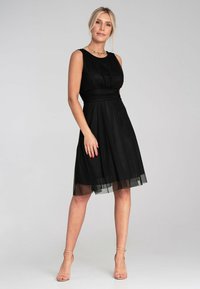 Schwarzes Kleid mit eng anliegendem Oberteil, gerafftem Taillenteil und weit auslaufendem Rock. Ein transparenter Überzug am Saum sorgt für Textur. Kombiniert mit nude-farbenen High Heels.