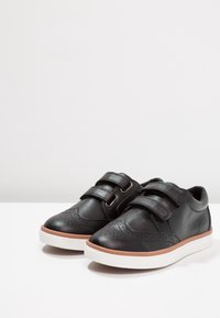 Baskets en cuir noir avec deux sangles Velcro, détails de brogue sur le bout, semelle en caoutchouc blanche et accents marron sur le bord.