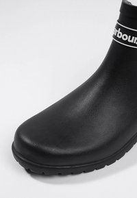 Botte en caoutchouc noire jusqu'à la cheville avec une texture lisse, un bout arrondi et une rayure blanche à logo autour du col. Semelle robuste et antidérapante.