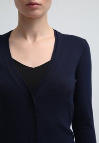 Cardigan blu navy in tessuto morbido, con scollo a V profondo, chiusura con bottoni e design aderente, che evidenzia una texture liscia.