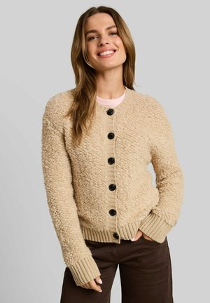 Jeune femme souriante, portant un cardigan beige texturé boutonné et un pantalon foncé, avec une main dans sa poche sur un fond uni.