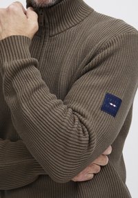 Barna bordás pulóver magas gallérral, texturált anyagból, bal karján kék, piros és fehér díszítéssel ellátott folttal.