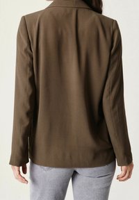 Hnědý blazer z hladkého materiálu, s upraveným střihem, špičatým klopem a dlouhými rukávy, kombinovaný se světle šedými džíny.