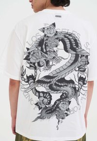 Persoon in een wit T-shirt met een groot zwart en grijs draak- en rozenontwerp op de achterkant, omgeven door cirkelvormige tekst.