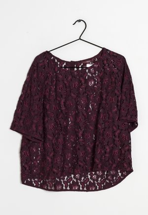 Blouse courte en dentelle violette avec motif floral accrochée à un cintre noir sur fond blanc.