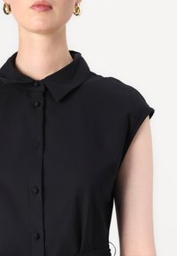 Camisa preta de manga curta com colarinho clássico; apresenta um fecho frontal com botões e um tecido liso e texturado.
