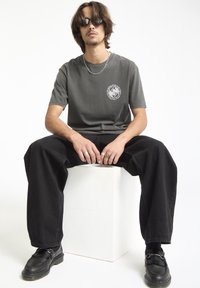 Grå bomulds t-shirt med rundt grafisk logo på brystet, kombineret med løse sorte denim bukser og sorte sko. Modellen bærer solbriller og ringe.