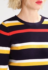 Pull en maille avec base marine et rayures horizontales rouges, jaunes et blanches. Conception à col rond, texture lisse et silhouette ajustée.