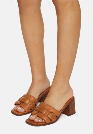 Brązowe skórzane sandały na słupku z przodem ozdobionym geometrycznymi wycięciami. Otwarty nosek, gładka faktura i drewniany obcas.