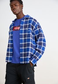 Blå og hvit rutete flannel-skjorte med svarte knapper over en blå grafisk t-skjorte, kombinert med svarte cargo-bukser med et logo-lapp.