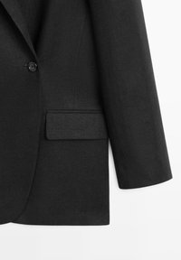 Zwarte op maat gemaakte blazer met een knoop aan de voorkant en een klepzak aan de rechterkant, getoond vanaf de onderkant van de romp en mouw tegen een witte achtergrond.