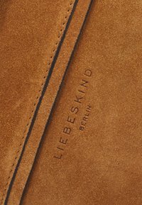 Tan-suede laukku, jossa on kohokuvioitu "LIEBESKIND BERLIN" -logo. Ominaisuuksiin kuuluu kontrastivärinen ommel ja pehmeä pinta, joka korostaa materiaalin laatua.